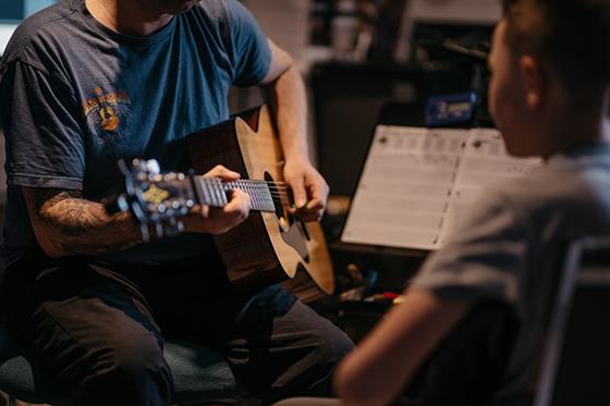 Photo of a man with tattoos teaching acoustic guitar to a child sitting across from him